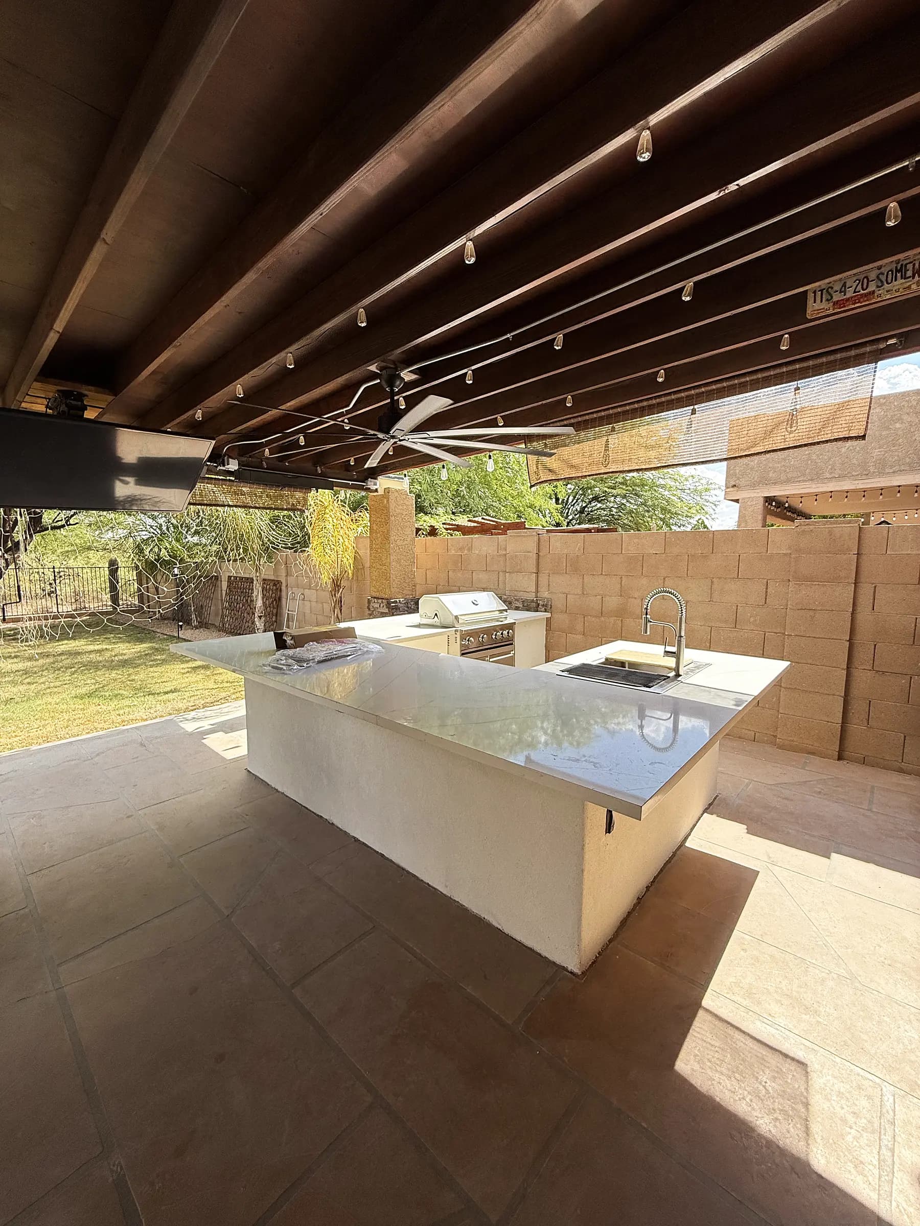 Long outdoor kitchen island with a sink and built-in grill under a beamed ramada strung with lights