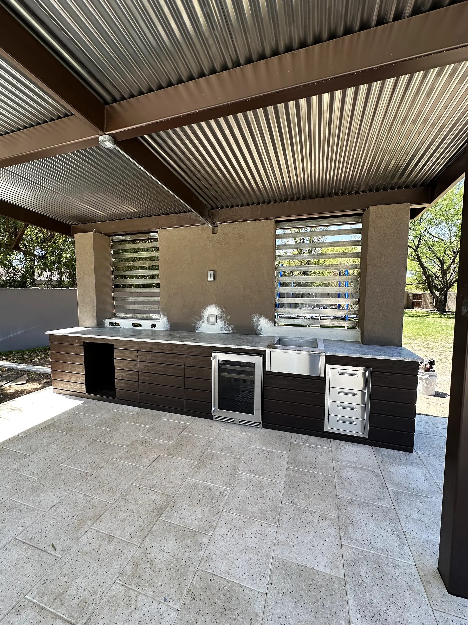 Modern outdoor kitchen with dark cabinetry, stainless sink and wine fridge under a corrugated metal shade structure